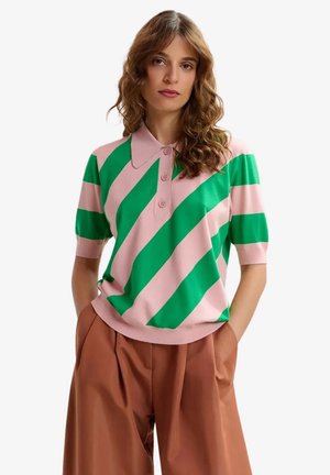 Donna con capelli mossi che indossa una polo a maniche corte a righe diagonali rosa e verde e pantaloni marroni a vita alta con le mani nelle tasche.