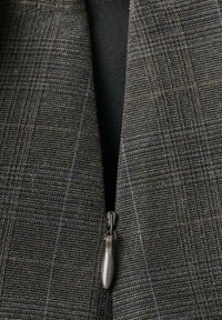 Chaqueta con cremallera y patrón de cuadros en tonos oscuros. Presenta un tirador de cremallera gris mate y tejido texturizado. El forro es negro.