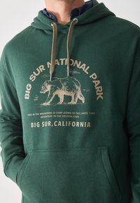 Sudadera verde con cordón, que presenta una ilustración de un oso y el texto "Parque Nacional Big Sur" en beige, material de algodón suave.