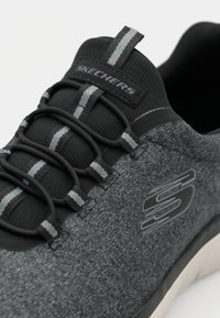 Musta urheilukenkä, jossa on teksturoitu harmaa päällinen, joustavat nauhat ja kumipohja. Sivussa on Skechers-logo.