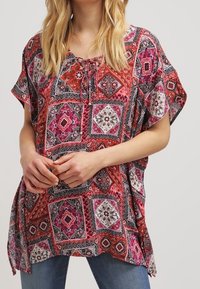 Tunique à manches courtes et ample en rose et rouge vifs, présentant un motif de patchwork géométrique avec des accents floraux et un détail à nouer au niveau du col.