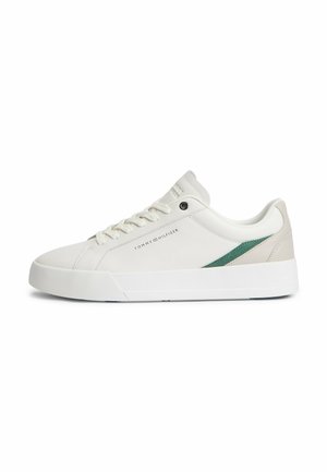 Witte lage sneaker met witte veters, groene zijstreep, beige hielaccent en "Tommy Hilfiger" logo aan de zijkant.
