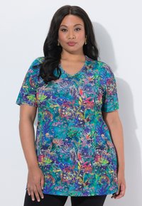 Kvinde med langt mørkt hår iført en farverig blomstret kortærmet tunika og sorte bukser, stående mod en hvid baggrund.