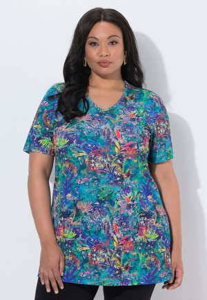 Kvinna med långt mörkt hår klädd i en färgglad blommönstrad kortärmad tunika och svarta byxor, stående mot en vit bakgrund.