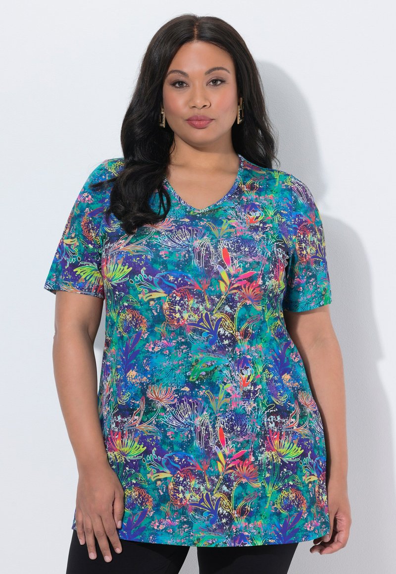 Kvinde med langt mørkt hår iført en farverig blomstret kortærmet tunika og sorte bukser, stående mod en hvid baggrund.