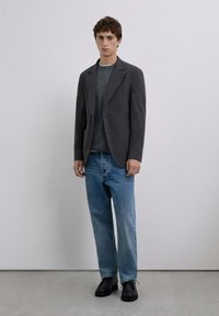 Blazer gris sobre un suéter gris oscuro, combinado con jeans rectos de mezclilla azul claro y zapatos negros. Tejido suave con un corte a medida.