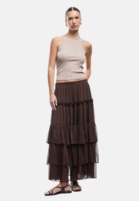 Beige geribbelde tanktop gecombineerd met een gelaagde, doorzichtige bruine maxi-jurk met gerafelde accenten en een elastische tailleband. Platte sandalen.