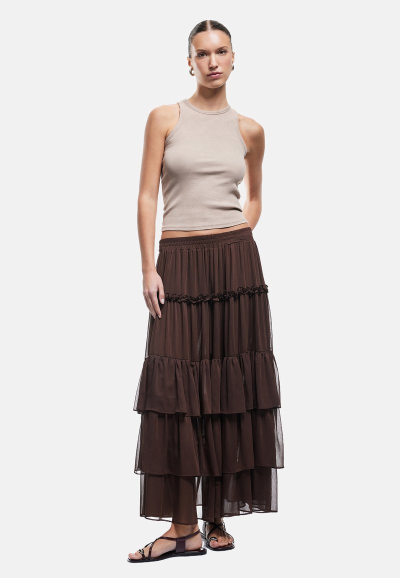 Beige geribbelde tanktop gecombineerd met een gelaagde, doorzichtige bruine maxi-jurk met gerafelde accenten en een elastische tailleband. Platte sandalen.