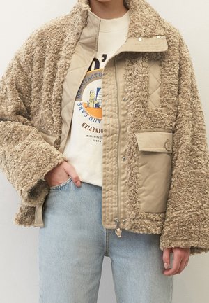 Veste en polaire sherpa beige avec panneau matelassé et poche à rabat, portée ouverte sur un sweat-shirt graphique et un jean bleu clair.