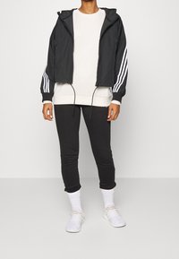 Giacca a vento nera con strisce bianche, indossata sopra un felpa grigia chiara e leggings neri. Scarpe sportive bianche con calzini bianchi.