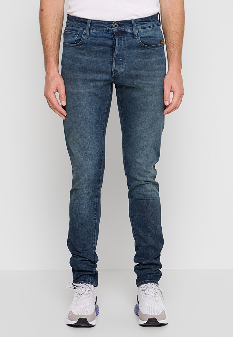 Homme portant un jeans slim fit bleu foncé et des chaussures de sport blanches, se tenant devant un fond clair et uni.