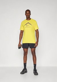 T-shirt de corrida amarela da Nike com logótipo cinzento, combinada com calções pretos. O modelo segura uma garrafa de água e usa ténis e meias pretos.