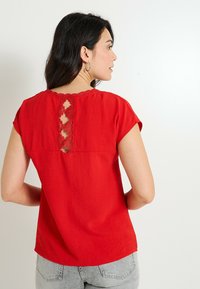 Haut rouge à manches courtes en tissu texturé, avec un détail de découpe en dentelle décorative dans le dos et une coupe décontractée.
