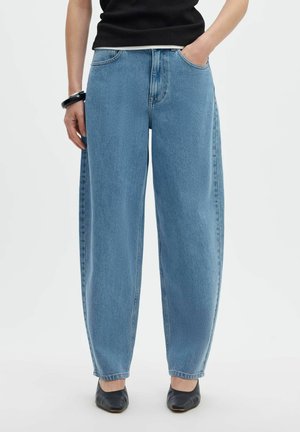Personne portant un jean bleu clair ample avec les mains dans les poches, un haut noir, un bracelet noir et des chaussures noires pointues.