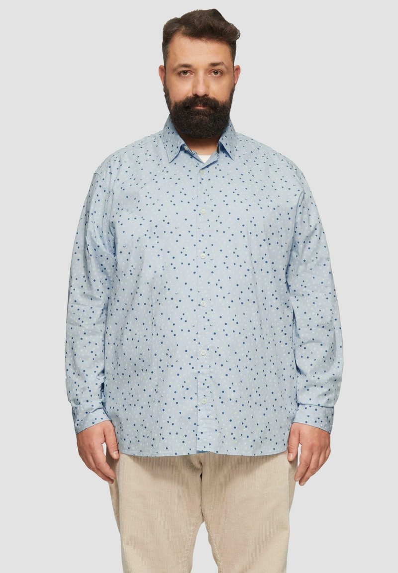 Lichtblauw overhemden met donkerblauwe stippen, lange mouwen en een kraag. Draagt met crèmekleurige corduroy broeken.