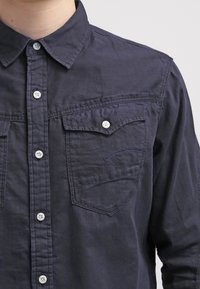 Dunkelblaue, knöpfbare Hemdjacke mit Kragen, zwei Brusttaschen und weißen Knöpfen. Der Stoff hat eine strukturierte Oberfläche und gestickte Akzente.