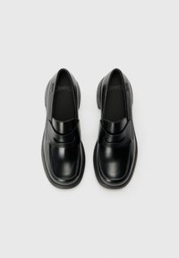 Juodos odinės slip-on tipo bateliai su lygiu paviršiumi, apvali noselė ir kontrastinga vidurinė dalis, pasižymintys minimalia siuvimo linija ir ženklinimu viduje.