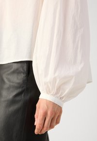 Gros plan sur la main et le poignet d'une personne portant une blouse blanche à manches longues avec des poignets froncés et un pantalon en cuir noir.