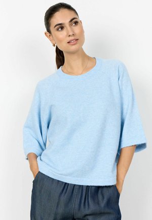 Kobieta o ciemnych włosach związanych w kucyk, ubrana w jasnoniebieski sweter z krótkim rękawem i ciemne spodnie, patrząca lekko na bok z rękami w kieszeniach.