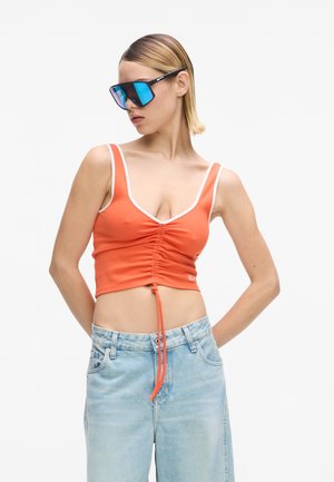 Femme portant des lunettes de soleil bleues réfléchissantes oversized, un crop top orange froncé avec des bordures blanches, et un jean taille haute bleu clair sur fond blanc.