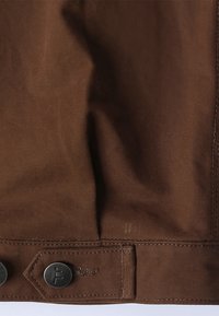 Καφέ ύφασμα με ορατή υφή, που διαθέτει μεταλλικά κουμπιά και ραφή κατά μήκος της άκρης. Ο σχεδιασμός περιλαμβάνει μια καπάκια με λεπτομέρεια επωνυμίας.