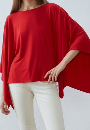 Femme portant un haut rouge ample à larges manches et un pantalon blanc ajusté, debout devant un fond clair uni.