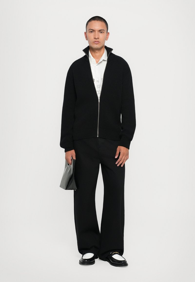 Veste zippée noire côtelée, chemise blanche à col en dessous, pantalon large noir, tenant une pochette noire et portant des chaussures noires et blanches.