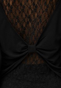 Crna majica s složenim crnim čipkastim detaljima, V-izrezom na leđima i mašnom u struku, koja naglašava teksturu i dizajnerske elemente.