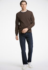 Brauner Rundhalspullover mit strukturiertem Finish, kombiniert mit dunkelblauen Jeans und weißen Sneakers. Das Model hat eine aufrechte Haltung und einen neutralen Gesichtsausdruck.