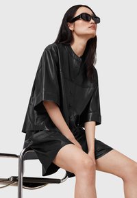 Black leather oversized shirt with short sleeves and button closure, paired with a matching leather skirt. Worn with black rectangular sunglasses.