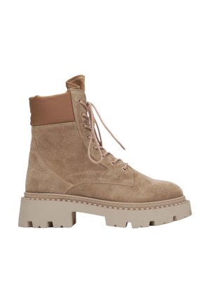 Beżowe zamszowe botki za kostkę z brązowym wyściółką, okrągłym noskiem i grubą beżową podeszwą o podwyższonej konstrukcji. Posiadają płaskie sznurowadła i boczne logo.