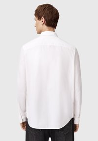 Camisa blanca de manga larga con cuello puntiagudo, textura ligera, detalle en la yugo trasero, dobladillo curvado y puños abotonados, llevada por una persona.