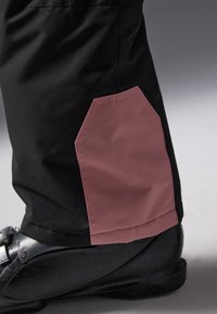 Black waterproof pants feature a pink accent patch at the lower leg, textured finish, and a slim fit design. Black footwear visible below.