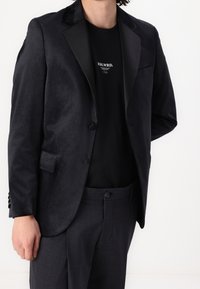 Man med en svart sammetblazer, svart mönstrad T-shirt och mörkgrå skräddarsydda byxor mot en vit bakgrund.