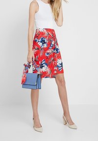 Mujer con una camiseta sin mangas blanca, falda envolvente Roja con flores, tacones beige, sosteniendo un bolso azul con una bufanda floral a juego en el asa.