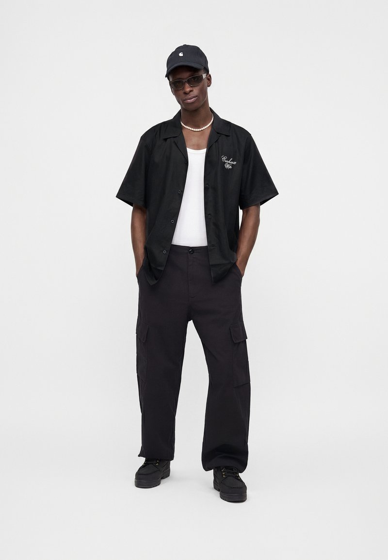 Giovane uomo con berretto nero, occhiali da sole, collana di perle, camicia nera a maniche corte sopra canottiera bianca, pantaloni cargo neri e scarpe nere.