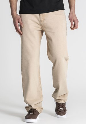 Person wearing beige loose-fit pants, brown sneakers with white soles, and a black shirt, standing on a white background.