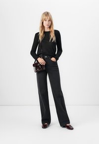 Blonde woman wearing black long-sleeve top and high-waisted wide-leg jeans, holding a small dark leather shoulder bag and wearing matching shoes.