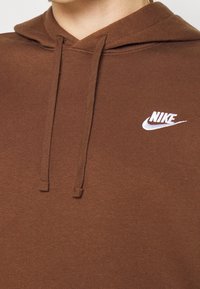 Hnědá mikina s kapucí vyrobená z měkkého materiálu, s bílým logem Nike na levé horní straně a šňůrkovým detailem u výstřihu.