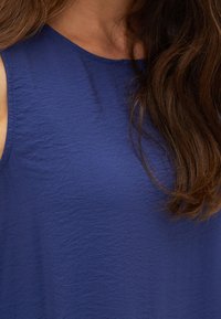 Hihaton sininen pusero, joka on valmistettu teksturoidusta kankaasta. Siinä on pyöreä pääntie ja rento istuvuus. Ei näkyviä kuvioita tai metalliosia.