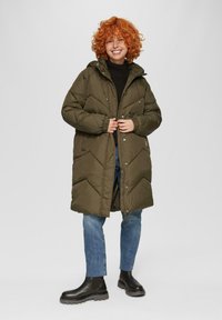 Olijfgroene pufferjas met een chevronpatroon, ritszakken, een capuchon en verstelbare manchetten. Draagt over een zwarte trui en jeans.