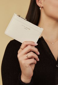 Cream leather wallet with a textured finish, zip closure, and embossed branding. Held by a hand wearing rings, against a neutral background.