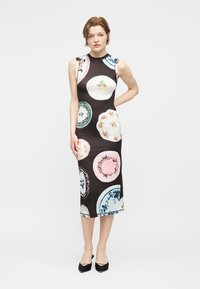 Γυναίκα που φοράει μαύρο μίντι φόρεμα χωρίς μανίκια με πολύχρωμα σχέδια σε στυλ ρετρό πιάτων, στέκεται με το ένα χέρι πίσω από την πλάτη και μαύρα τακούνια.