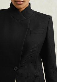 Must mustrindiga bleiser kõrge kaelusega, tekstureeritud kangast, must ühekordne nööp ja tasku vasakul rinnal. Diagonaalne ribakujundus.