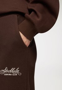 Brązowy sweatshirt z dzianinowymi mankietami, z boczną kieszenią i haftowanym logo "Stockholm Leisure Club" w białym kolorze. Miękka tekstura, luźny design.