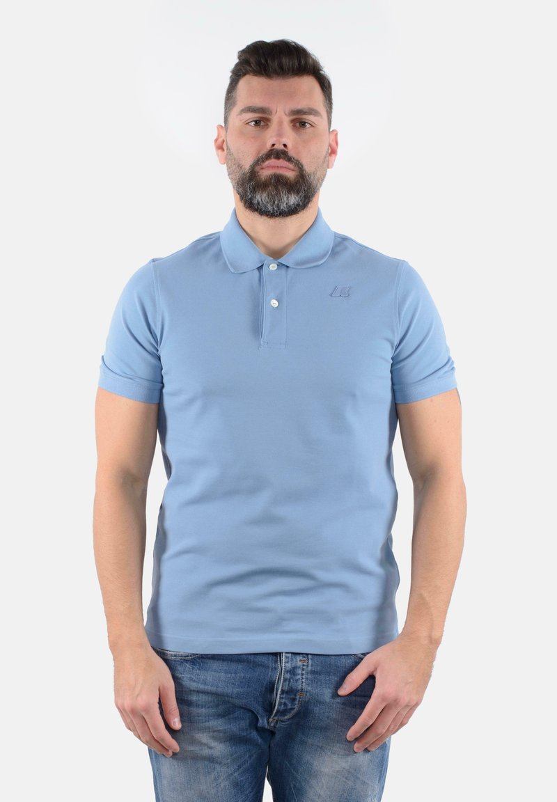 Polo azzurro chiaro con maniche corte, tre bottoni e un logo strutturato sul petto. Abbinato a jeans in denim sbiaditi.