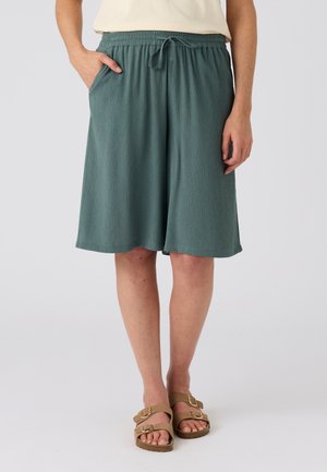 Vrouw draagt losse groene knielange shorts met een elastische tailleband en trekkoord, gecombineerd met beige sandalen met gespen.