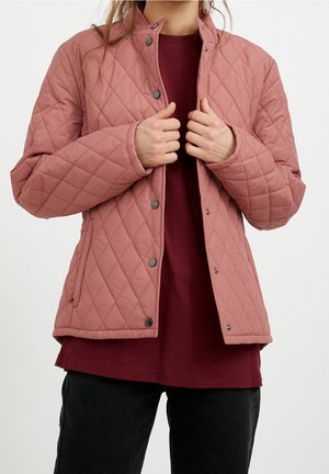 Persoon die een stoffige roze doorgestikte jas dichtdoet over een bordeauxrode blouse, zwarte broek draagt, getoond van nek tot midden dijbeen.