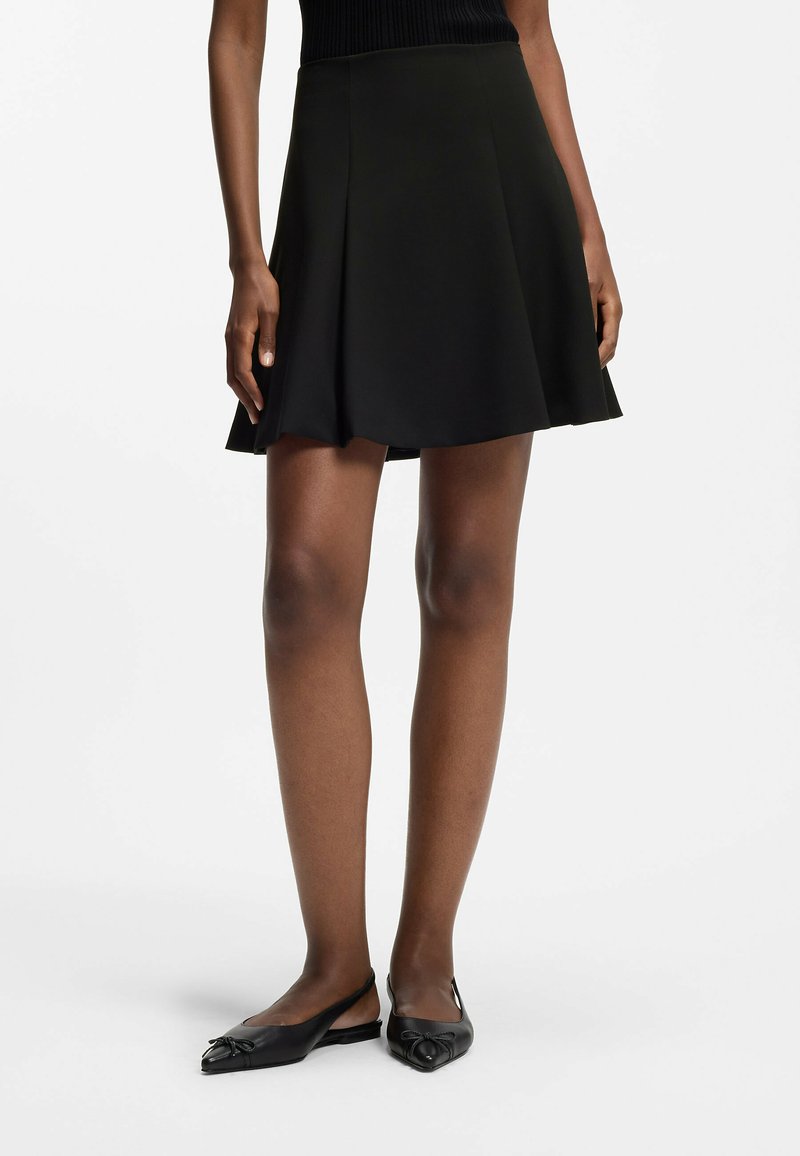 Frau trägt einen schwarzen A-Linien-Rock, ein schwarzes ärmelloses Oberteil und schwarze Slingback-Ballerinas mit spitzer Spitze und Schleifendetails.