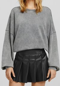 Grauer übergroßer Pullover mit gerippten Bündchen, kombiniert mit einem schwarzen, plissierten Rock aus Kunstleder mit figurbetontem Bund und dezentem Glanz.